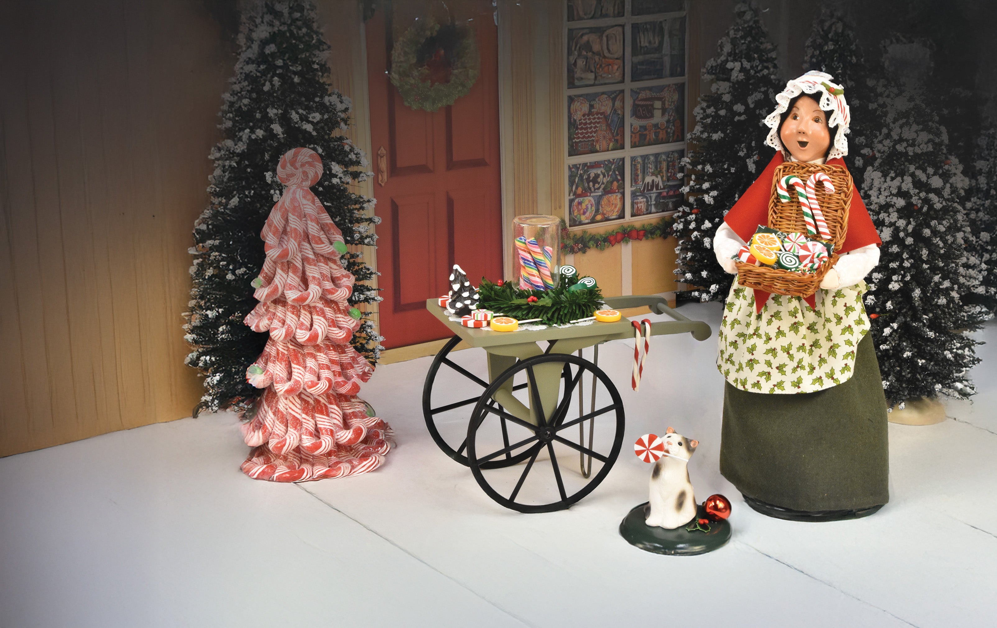 Photo of a Byers' Choice caroling Christmas  figurine holding a basket of Christmas candy with a grey and white cat at her feet with a candy cane in it's mouth and a candy cart prop at her side.